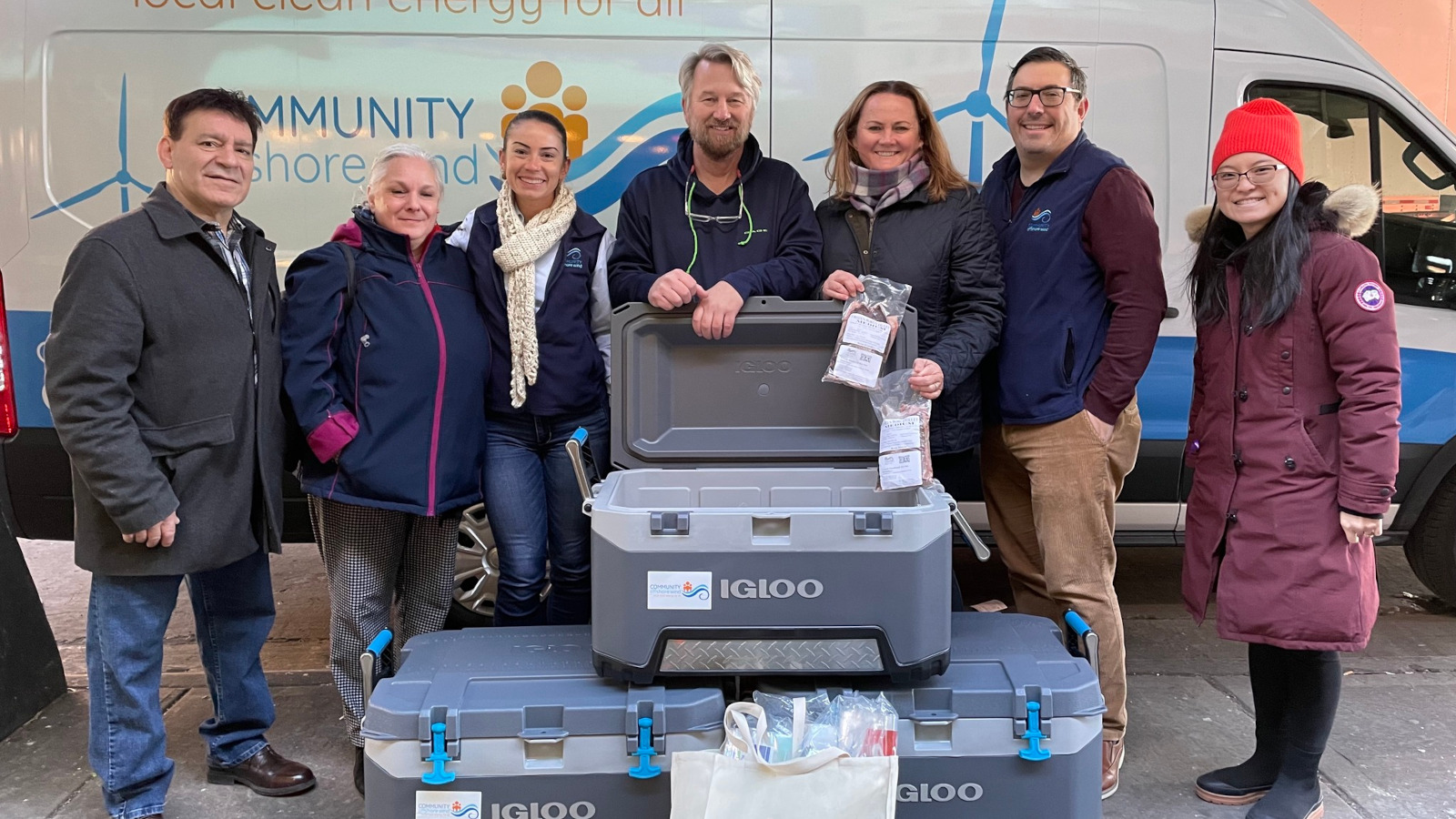 A group of people poses with open grey Igloo coolers. A van in the background reads 'COMMUNITY'.