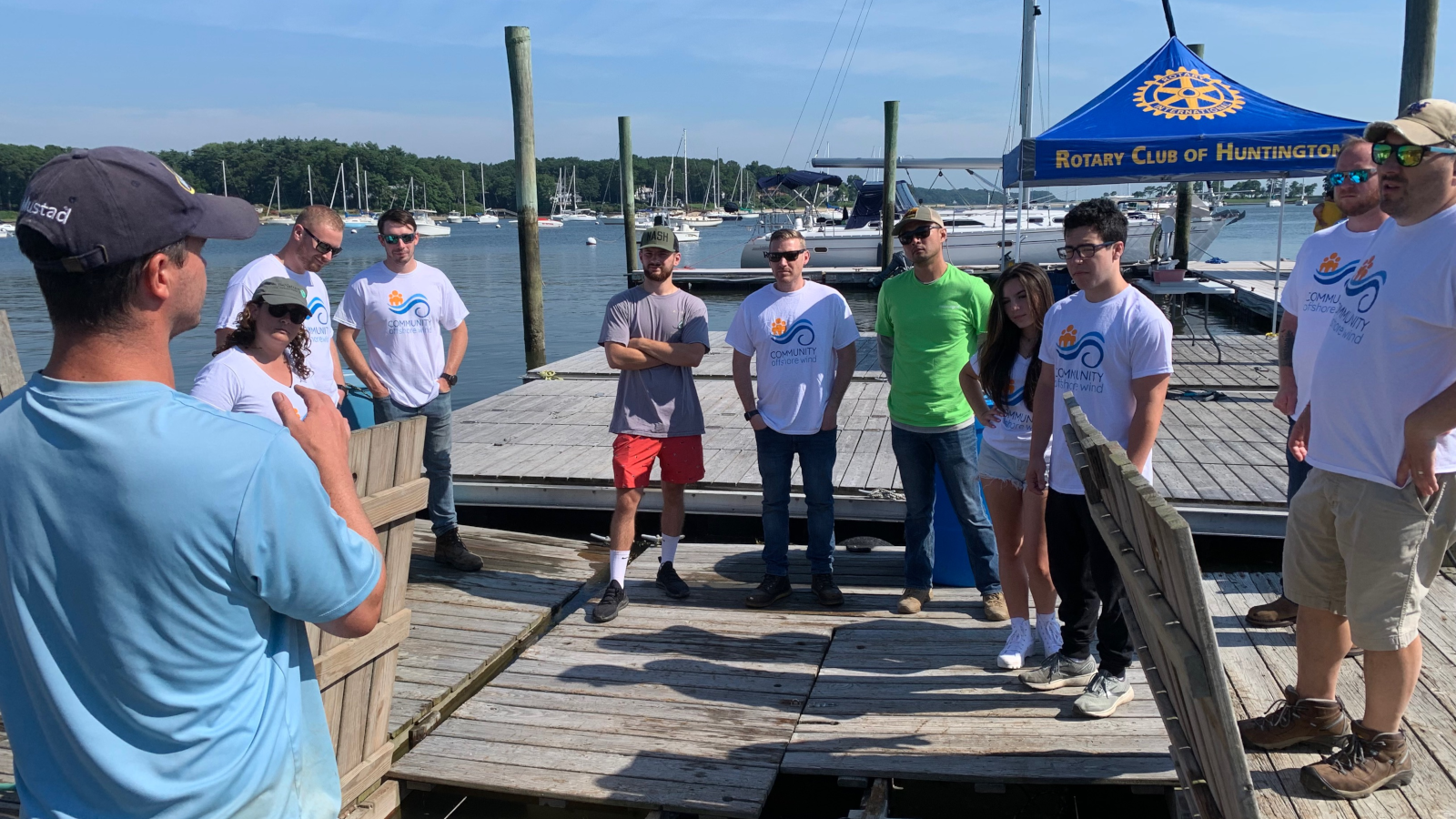 A group of volunteers listens to a speaker on a dock near a marina, with sailboats and a Rotary Club tent in the background.