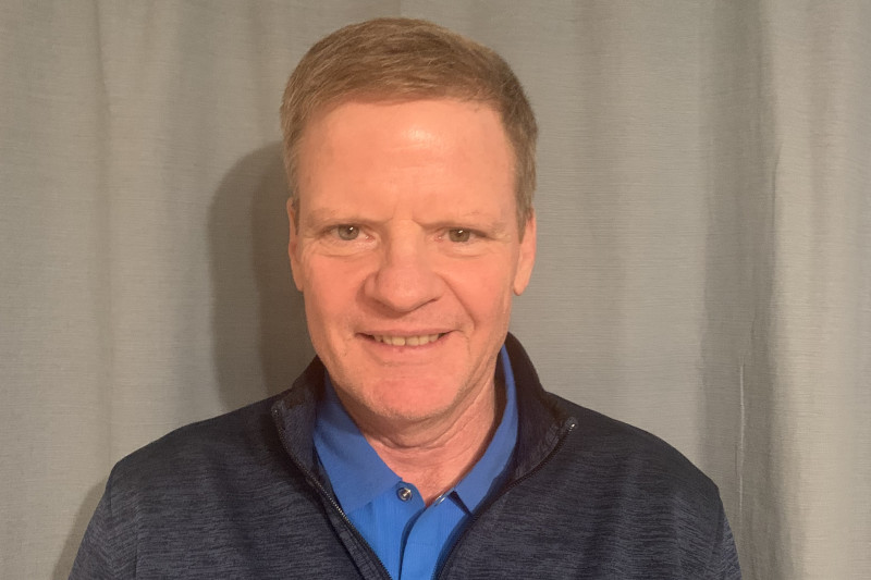A man wearing a navy blue jacket over a blue shirt stands against a grey backdrop.