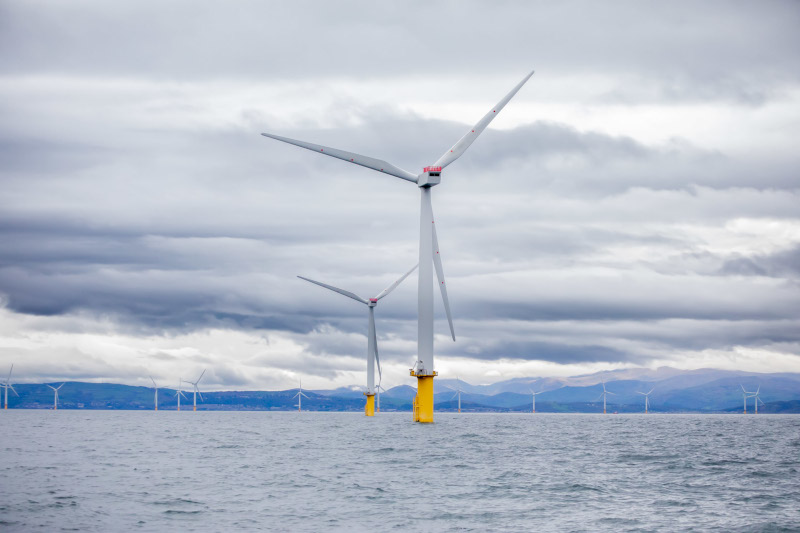 Three wind turbines stand on yellow pillars in a calm sea, under a grey sky with rolling clouds and distant mountains.