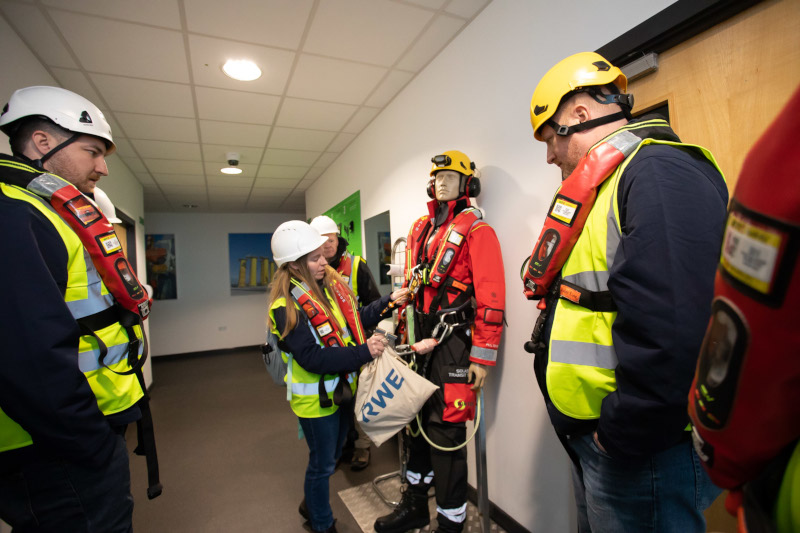 A group of workers in safety gear, including helmets and harnesses, examines a mannequin dressed in rescue clothing.