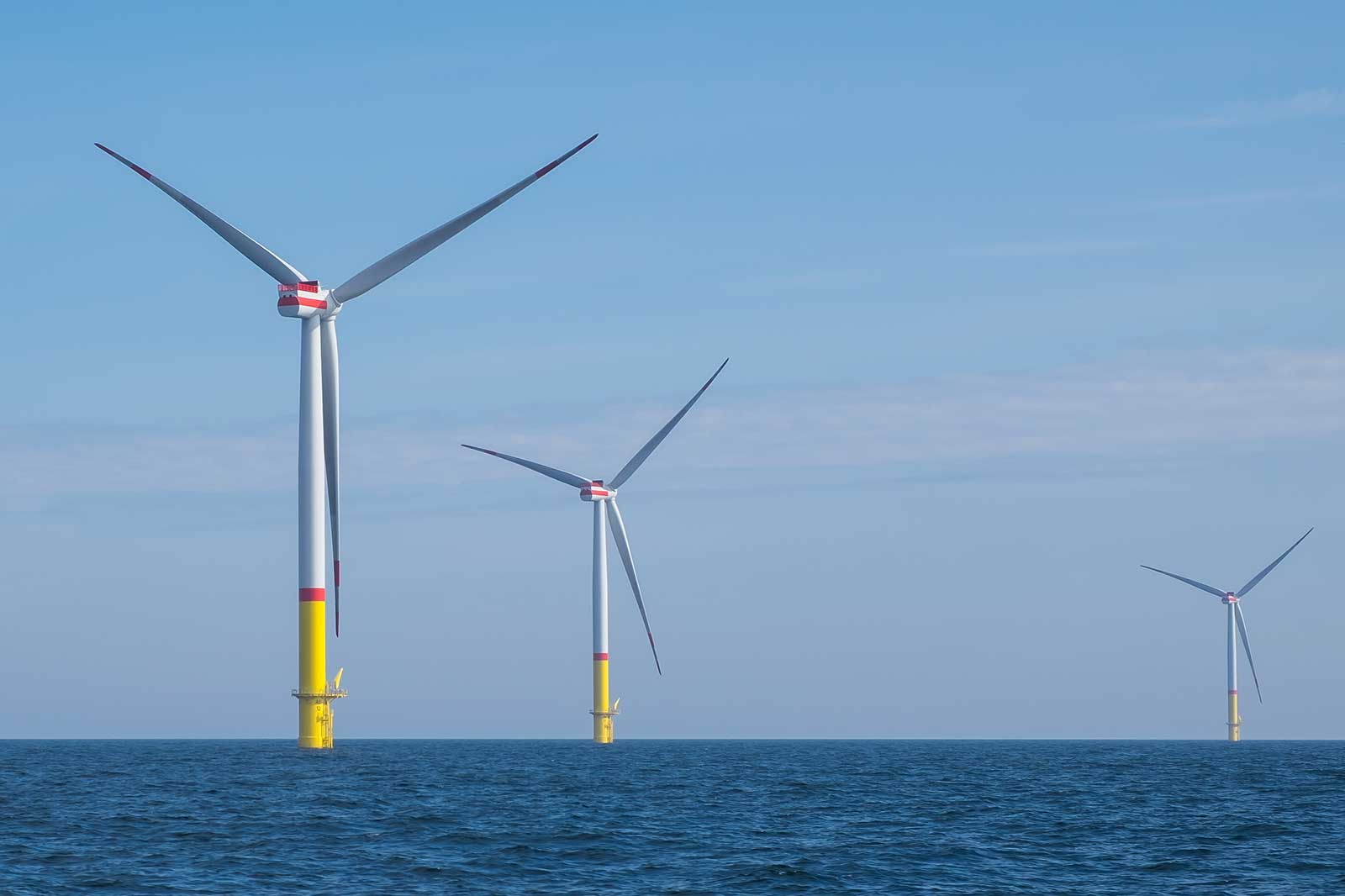 Three large offshore wind turbines stand in calm blue waters under a clear sky with soft clouds.