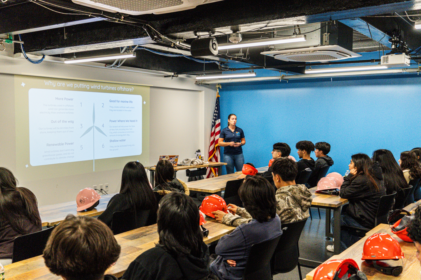 A presenter discusses offshore wind turbines with an audience in a classroom, with safety helmets on tables and an American flag in view.