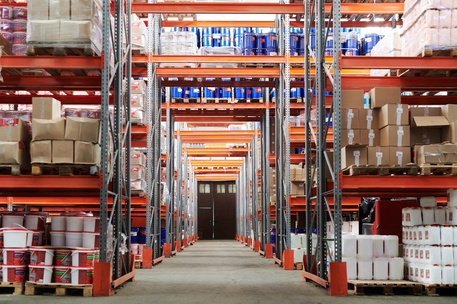 A spacious warehouse aisle with stacked shelves filled with boxes and containers, leading to a wide wooden door at the end.