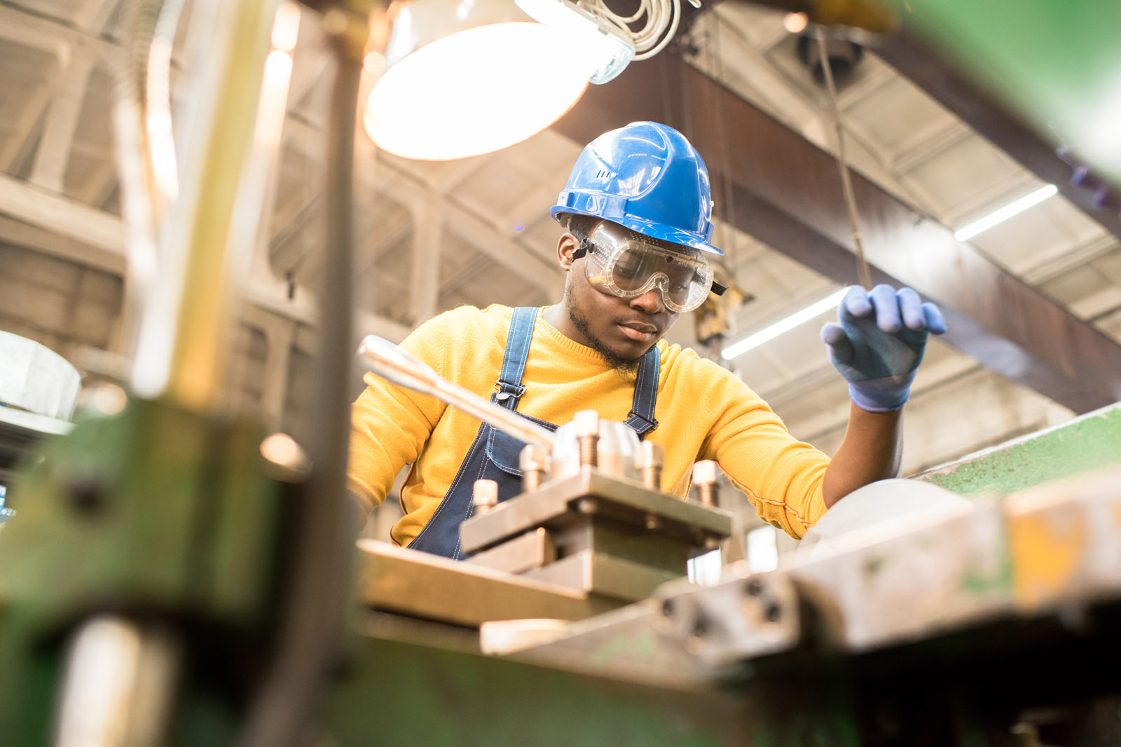 A worker in a blue helmet and gloves operates machinery in a workshop, focused on their task amidst industrial surroundings.